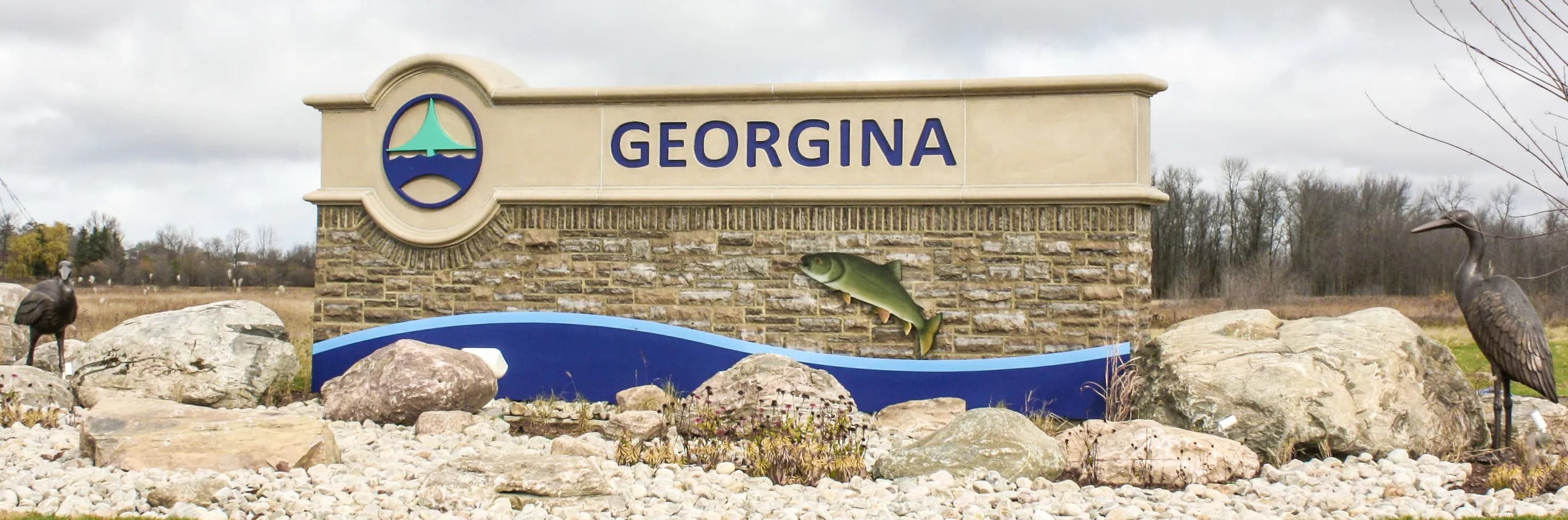Stone Town of Georgina welcome sign with a leaping fish and heron sculptures under a cloudy sky