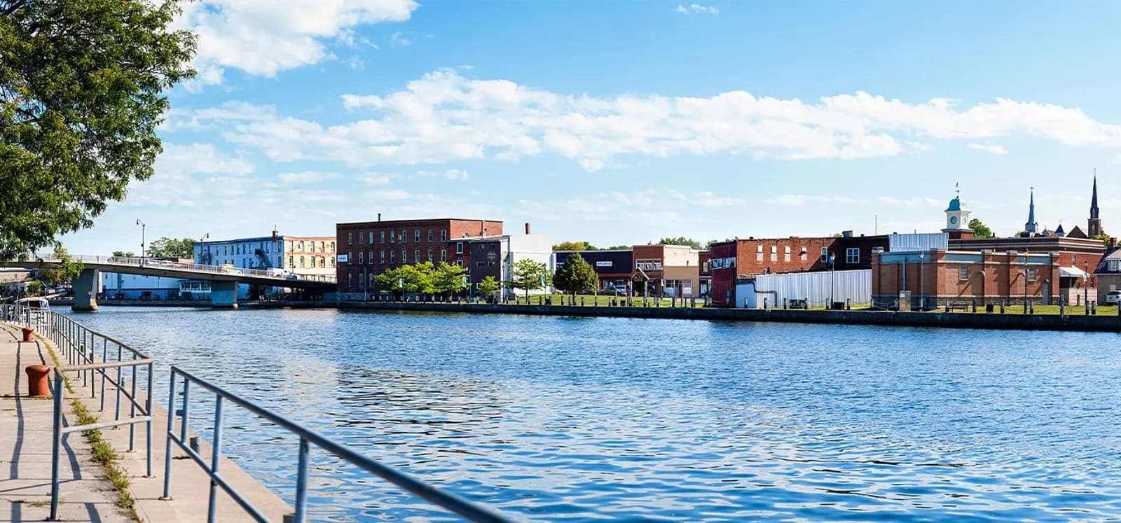 The Trent River flowing past Campbellford's downtown waterfront, with brick main-street buildings, a bridge and church spires along the far bank