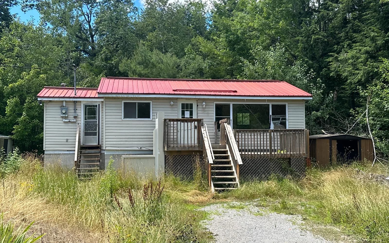 Grey home with a red metal roof and deck on an overgrown lot in Quinte West (Trenton), Ontario